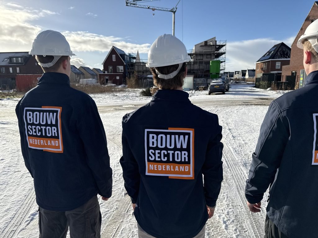 Drie vakmensen van Bouwsector Nederland met witte helm en bedrijfsjas kijken naar huizen in aanbouw in de sneeuw